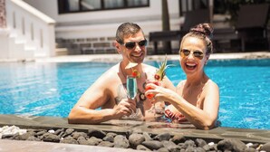 A couple in a swimming pool with drinks, surrounded by rocks and a building in the background. - TTC Hotel – Hoi An (Da Nang)