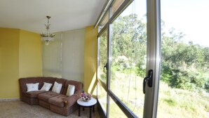 A room with a brown sectional sofa, a small round table with a vase, and a large window offering a view of greenery. - Hotel Galicia (Poio)