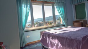 A bedroom with a bed, a window with curtains, a television, and a view of buildings and mountains. - Hotel Galicia (Poio)