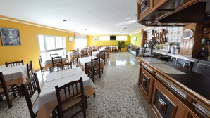 A dining area with multiple tables and chairs, a kitchen area with wooden cabinets and a black countertop, and a yellow wall with a window. - Hotel Galicia (Poio)