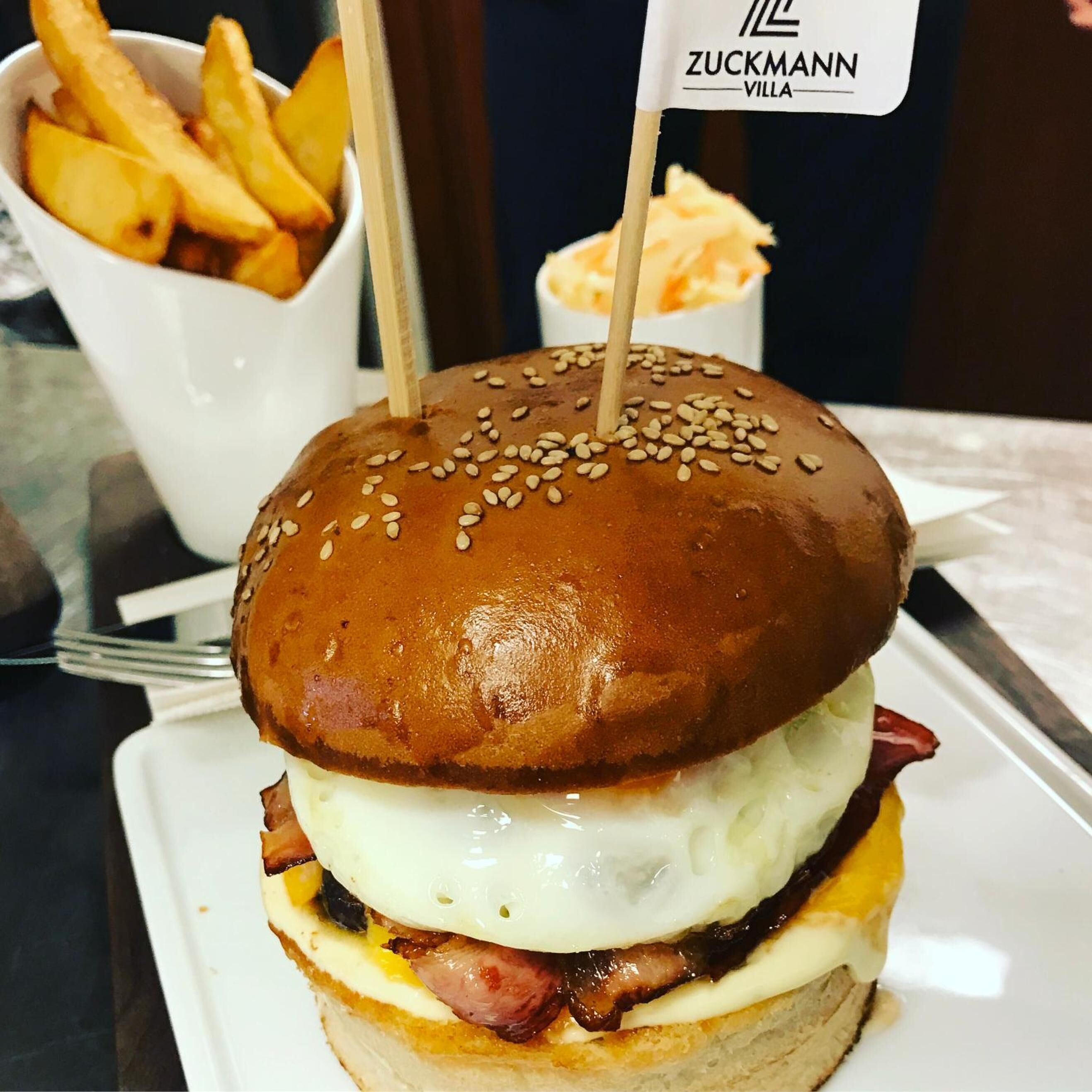 A sandwich with a sesame seed bun, cheese, bacon, and a fried egg, accompanied by fries and a small container of sauce.