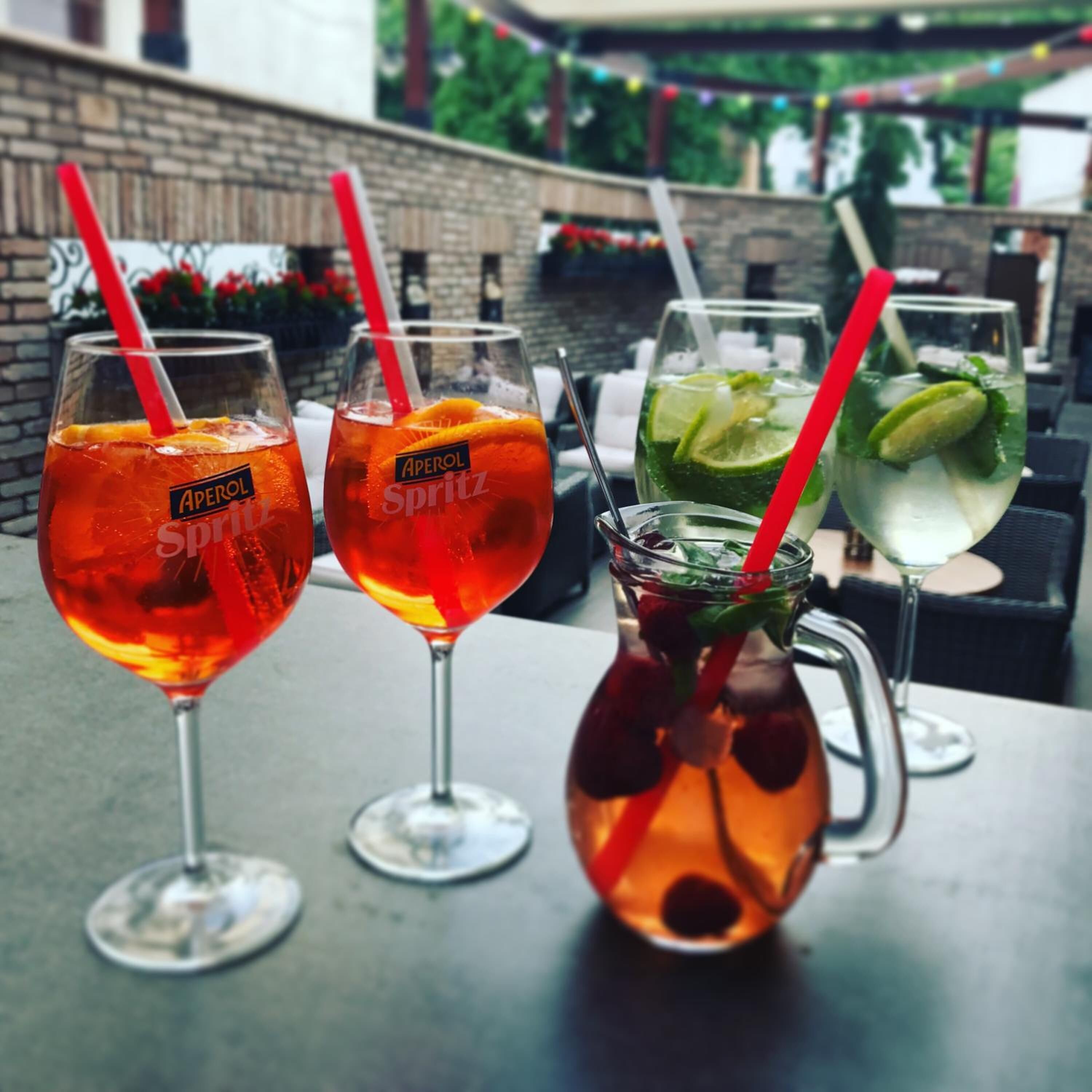 Drei Aperol Spritz und ein Krug mit einem roten Strohhalm stehen auf einem Tisch im Freien.