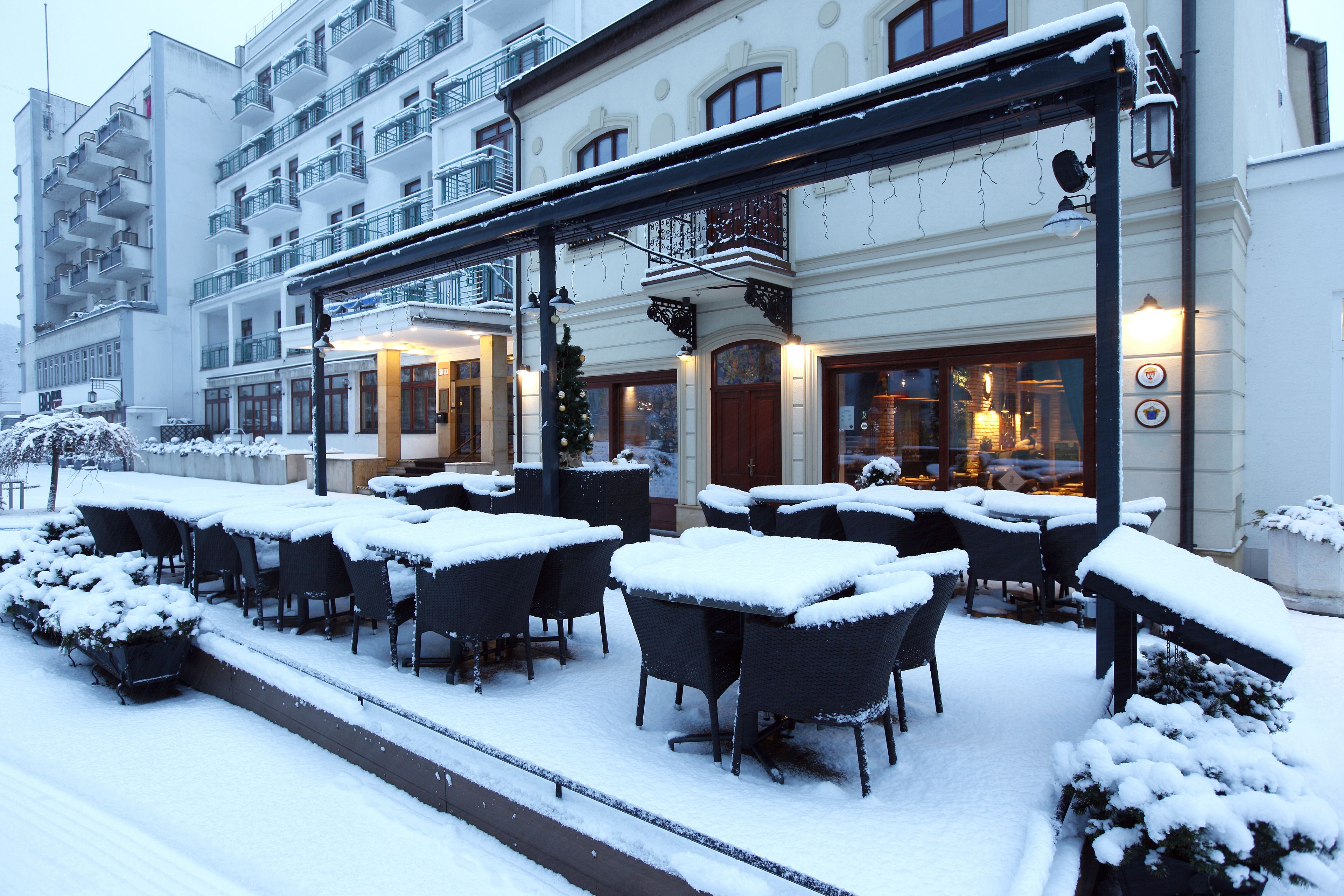A snow-covered outdoor seating area with tables and chairs, a building with multiple balconies, and a Christmas tree.