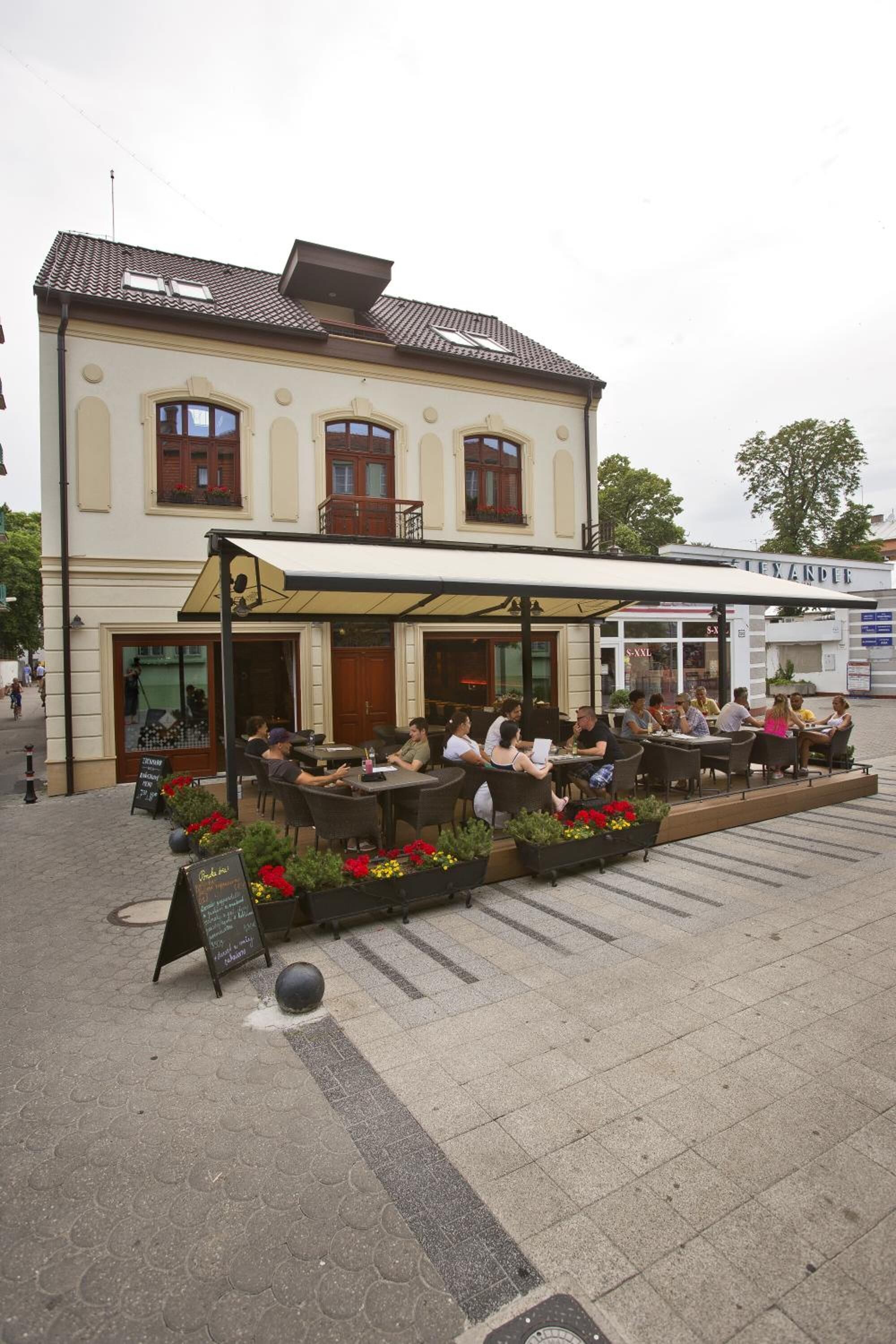 An outdoor cafe with tables and chairs, a building with a balcony, and a signboard.