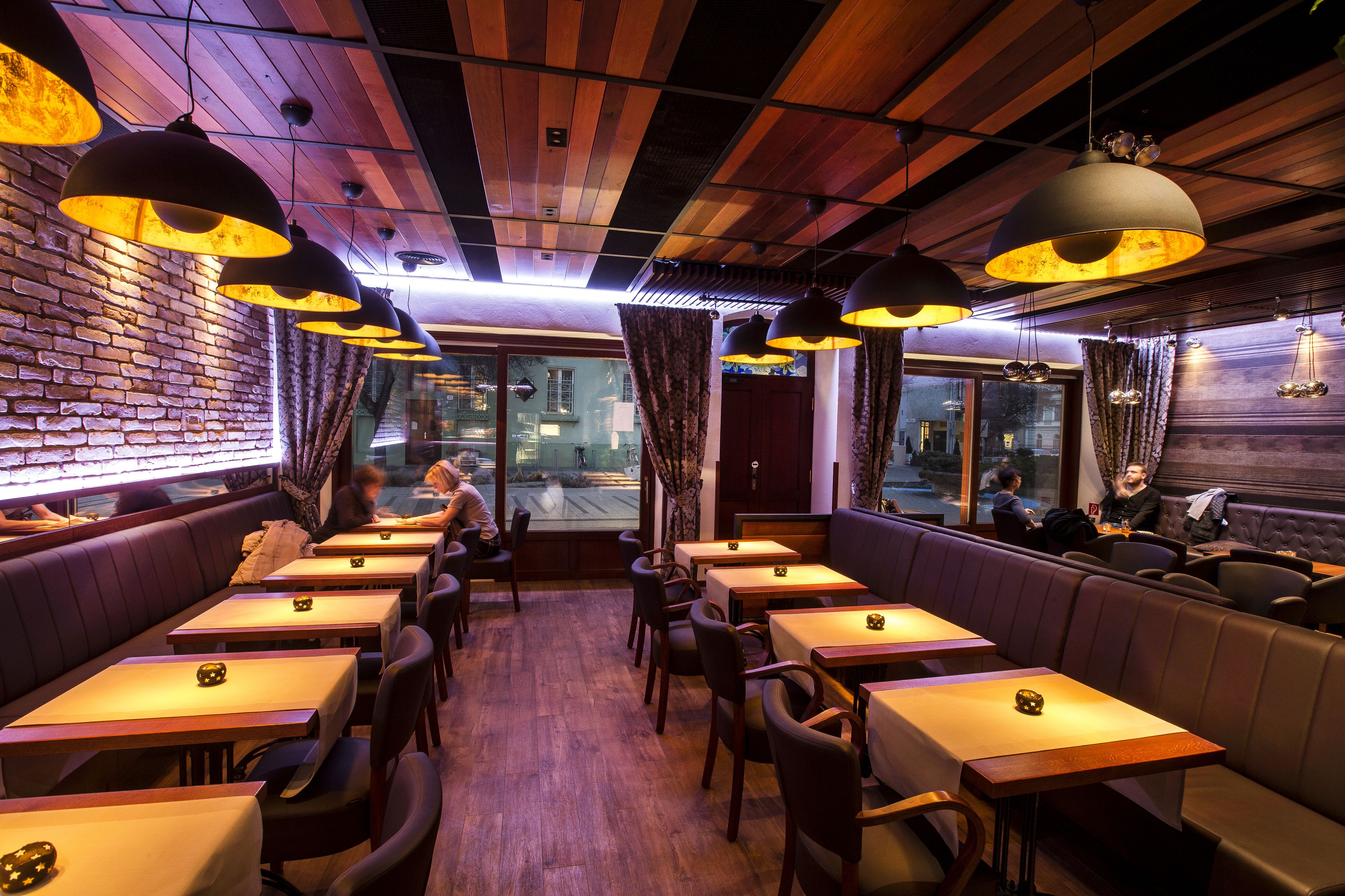 Ein Restaurant mit Holzbalkendecke, Pendelleuchten und einer Backsteinwand. Tische und Sitzecken laden zum Essen ein.