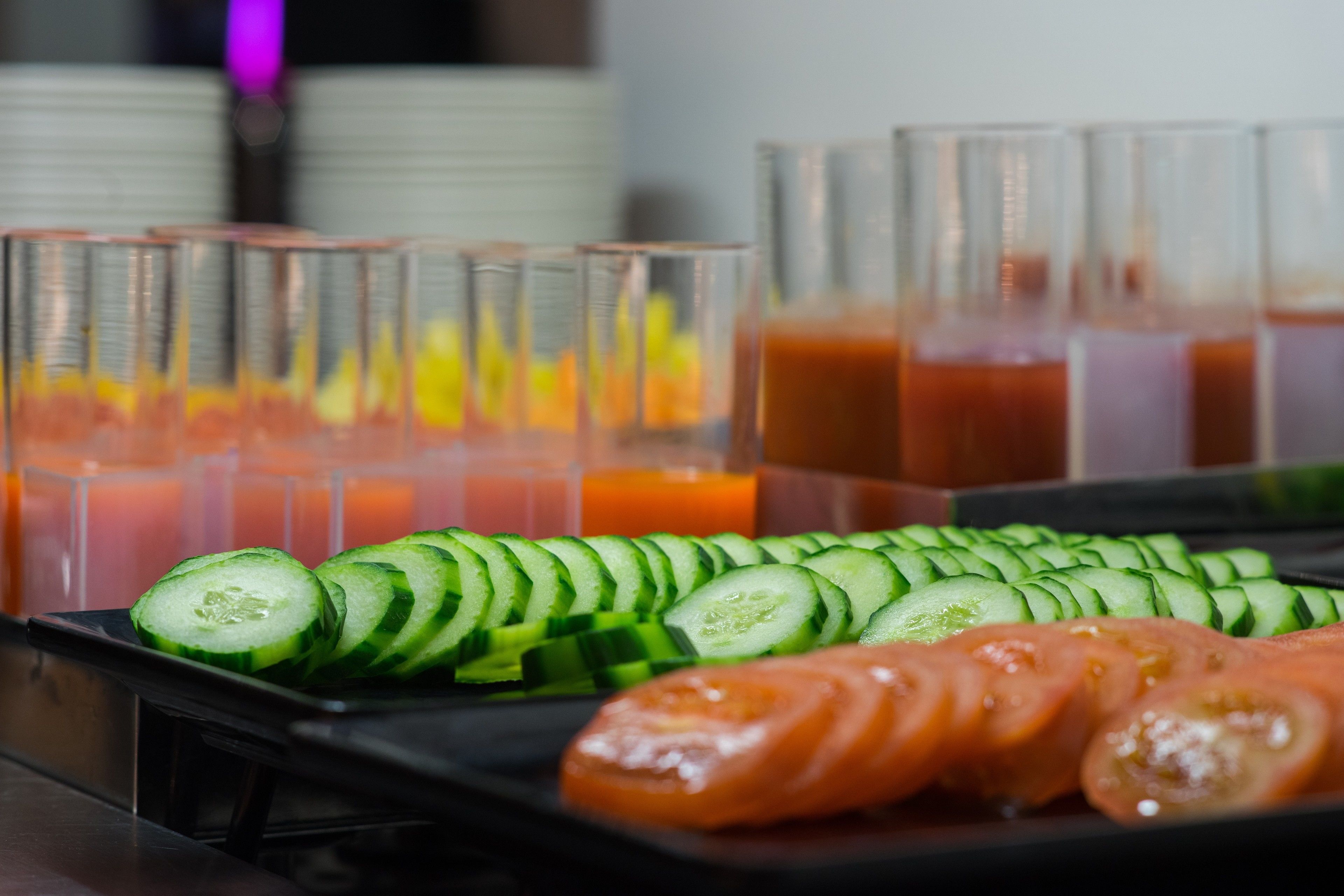 A tray of sliced cucumbers and tomatoes on a black surface with several glasses of juice in the background.