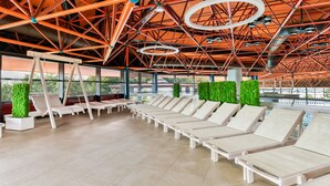 Un espace intérieur lumineux, baigné de soleil, avec des fauteuils blancs disposés en rangées, un plafond haut orné d’une structure métallique géométrique, et une vue sur la verdure à l’extérieur.