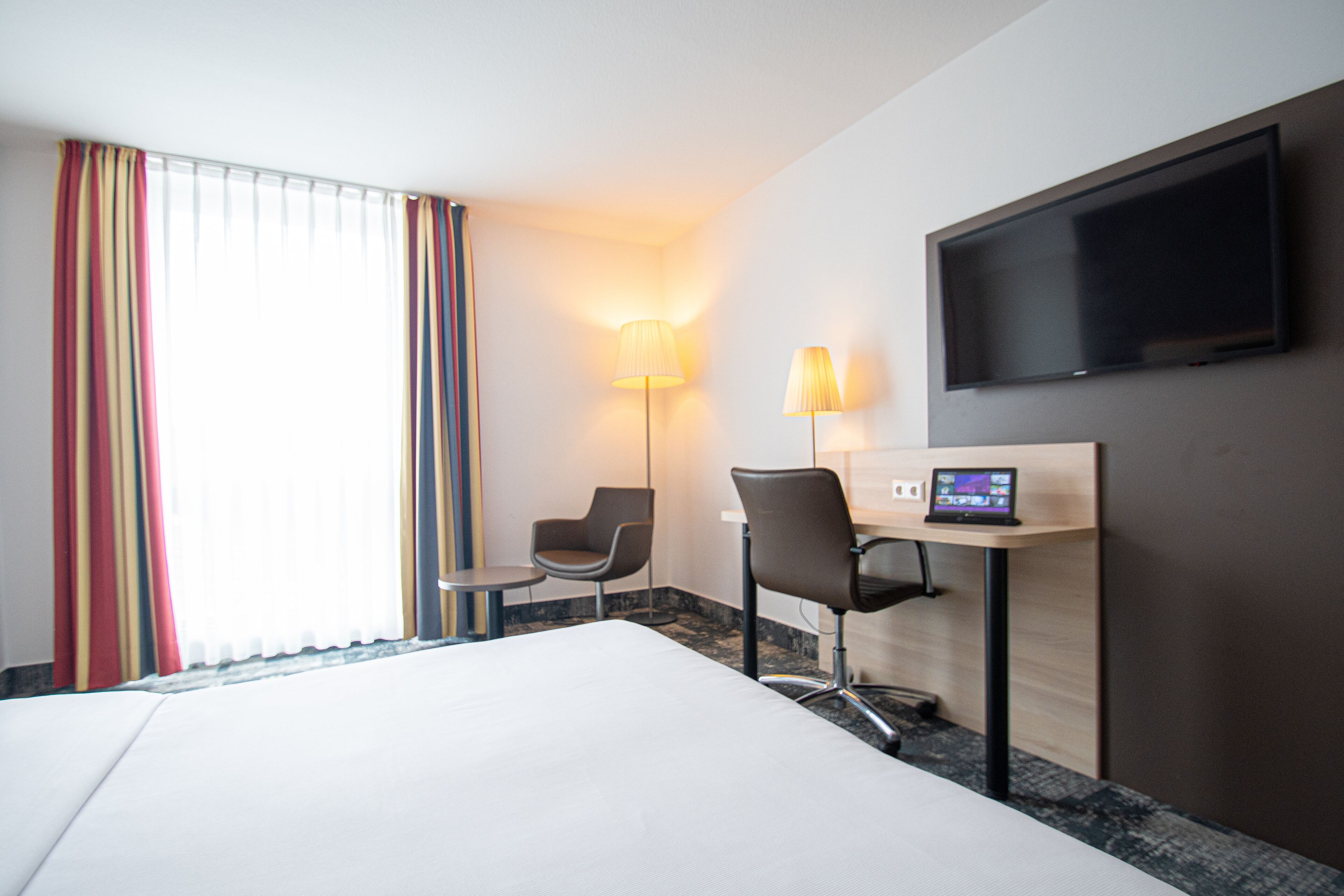 Une chambre d’hôtel comprenant un lit, un bureau avec un ordinateur, deux chaises, une télévision et une lampe.