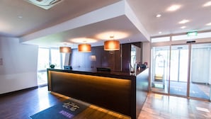 Un espace d’accueil d’hôtel avec un comptoir de réception en bois, des luminaires suspendus et une entrée avec une porte en verre.