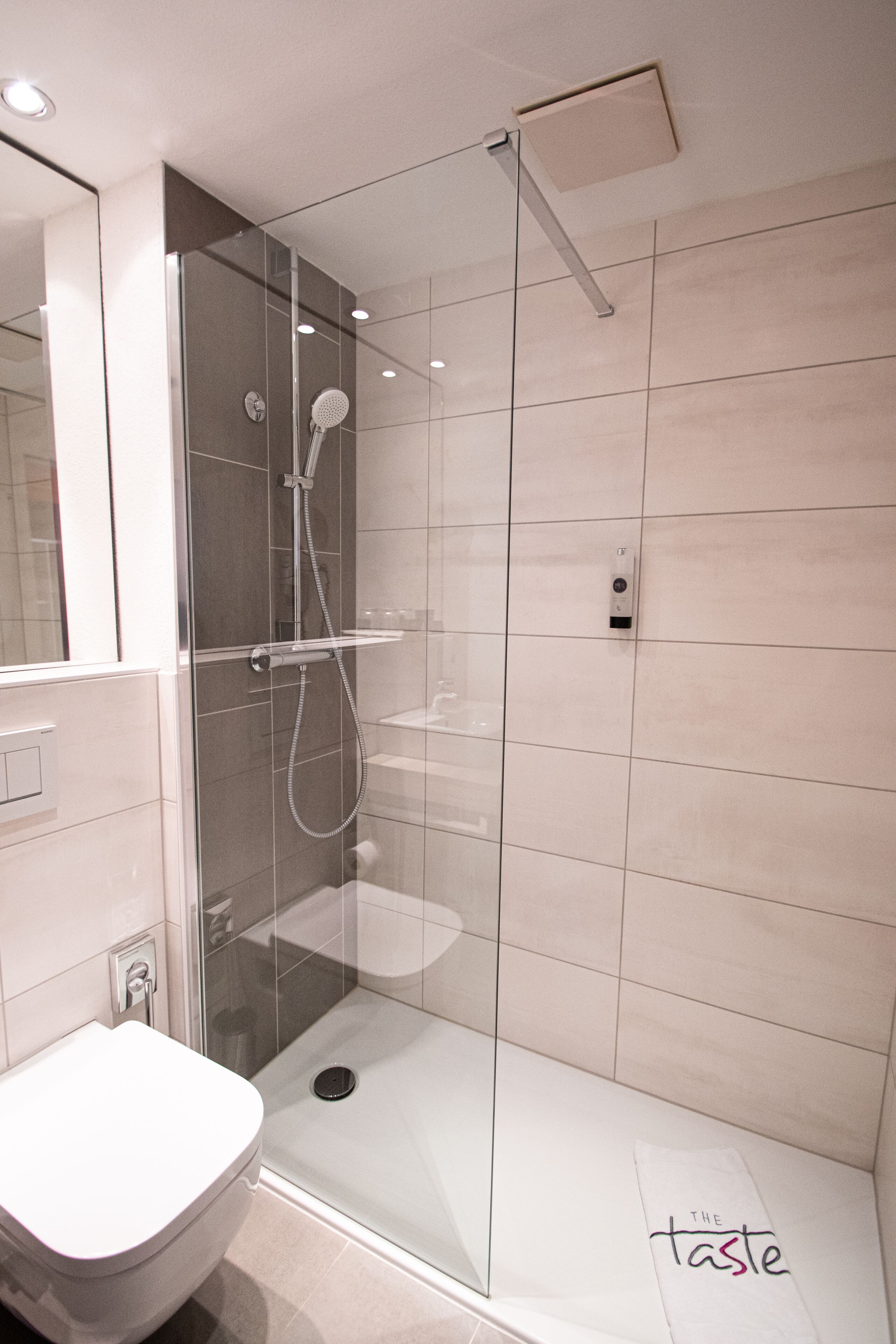 Une salle de bain moderne avec une douche à parois de verre, des toilettes blanches et un porte-serviettes sur lequel est posée une serviette portant l'inscription « The Taste ».