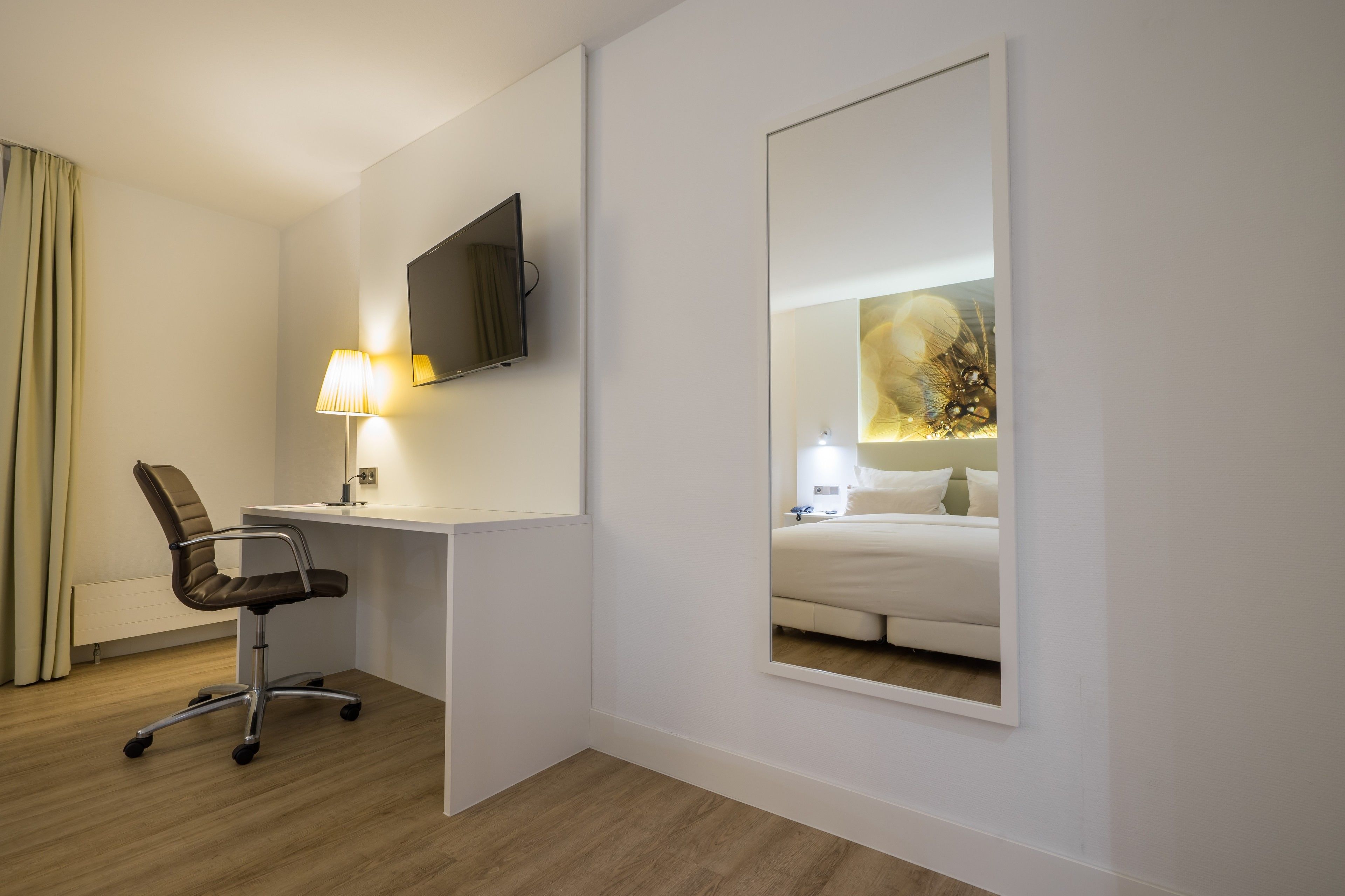 A hotel room with a desk, chair, TV, and a large mirror reflecting a bed and nightstand.