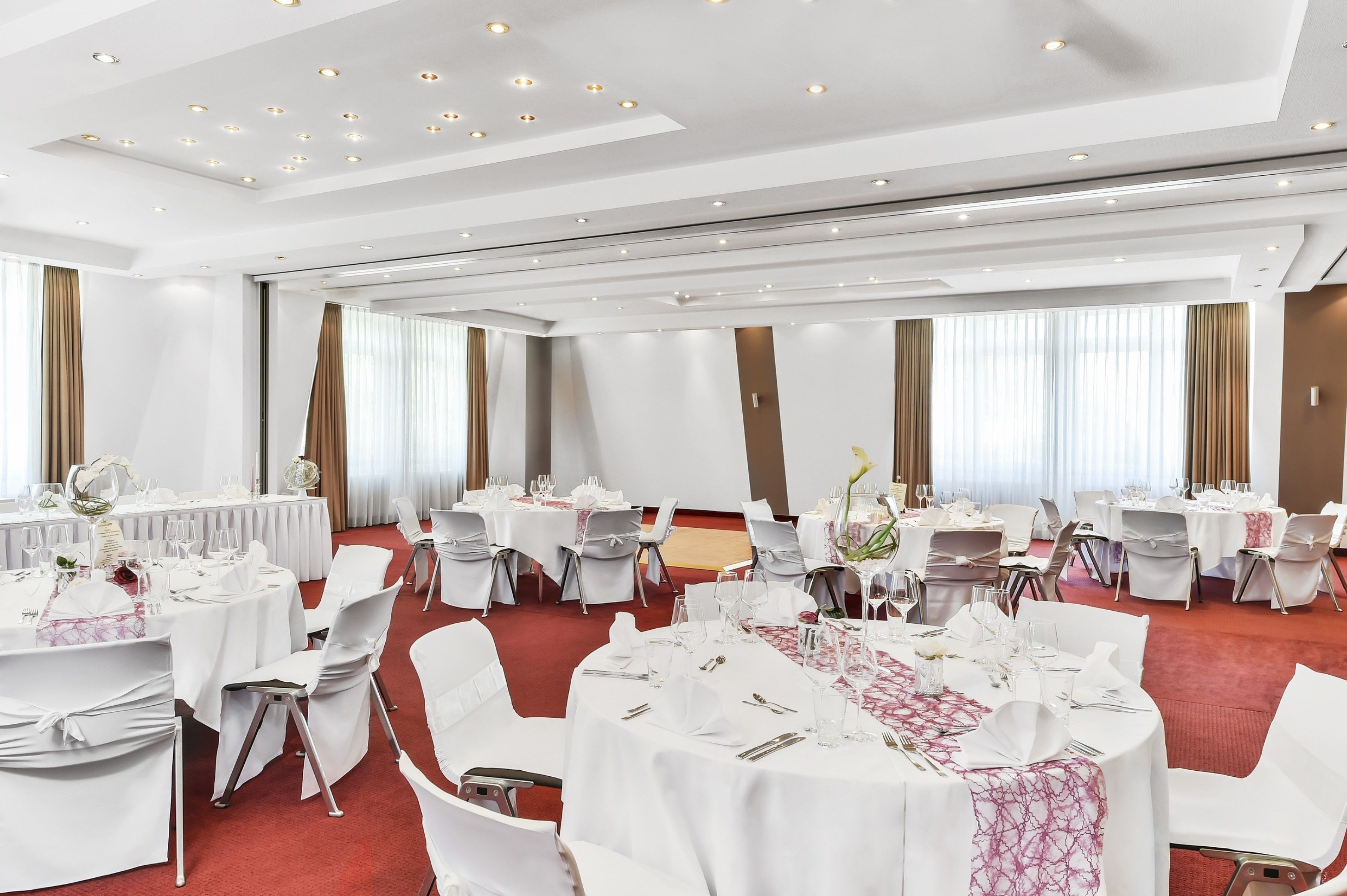 Un salón de banquetes con mesas redondas dispuestas para el servicio de comidas, sillas blancas y una alfombra roja.