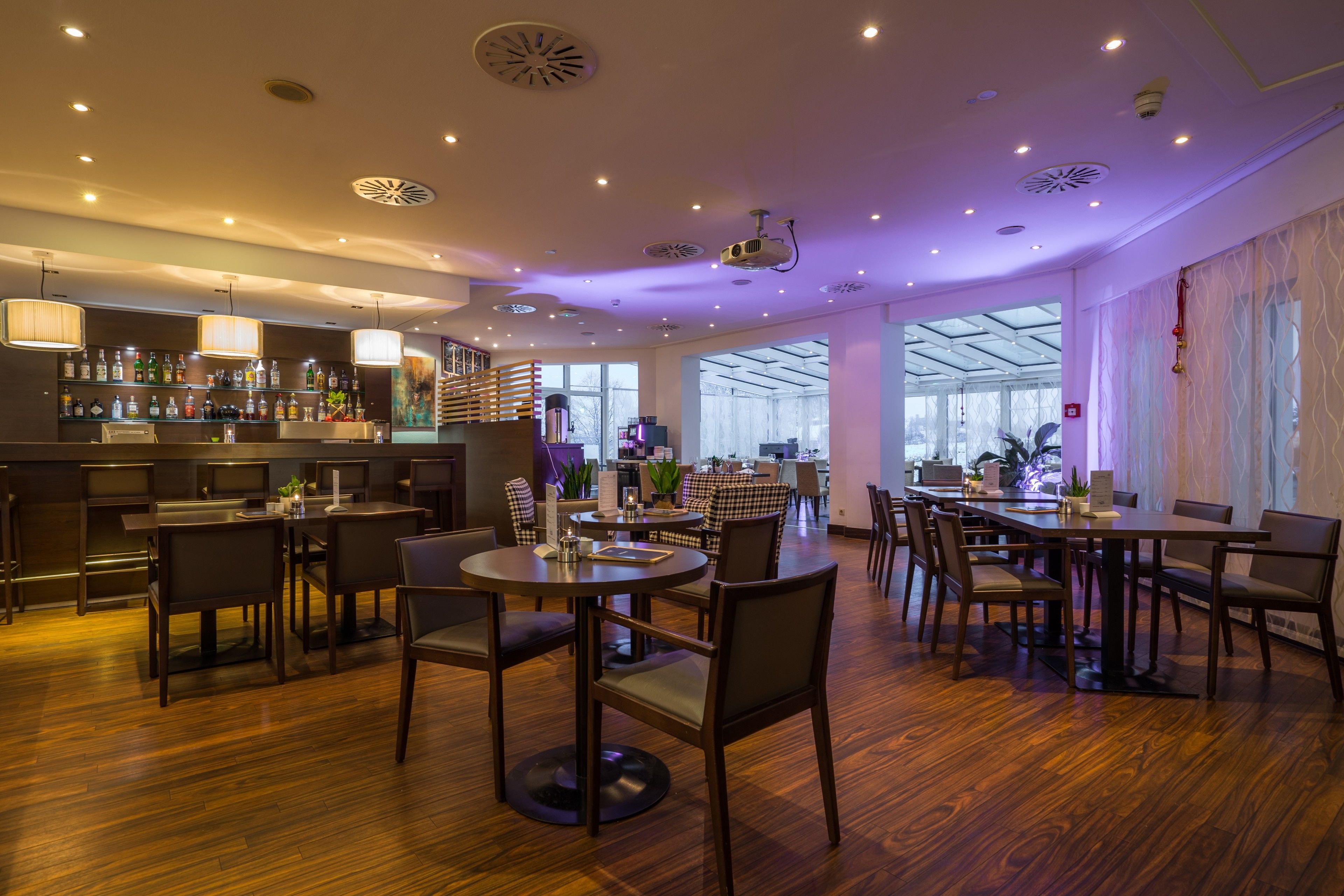 Un restaurante moderno con suelo de madera, mesas redondas y sillas. Cuenta con una zona de bar con estanterías llenas de botellas y una escalera que sube a un nivel superior.