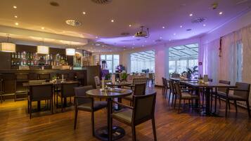 Un restaurante moderno con suelo de madera, mesas redondas y sillas. Cuenta con una zona de bar con estanterías llenas de botellas y una escalera que sube a un nivel superior.