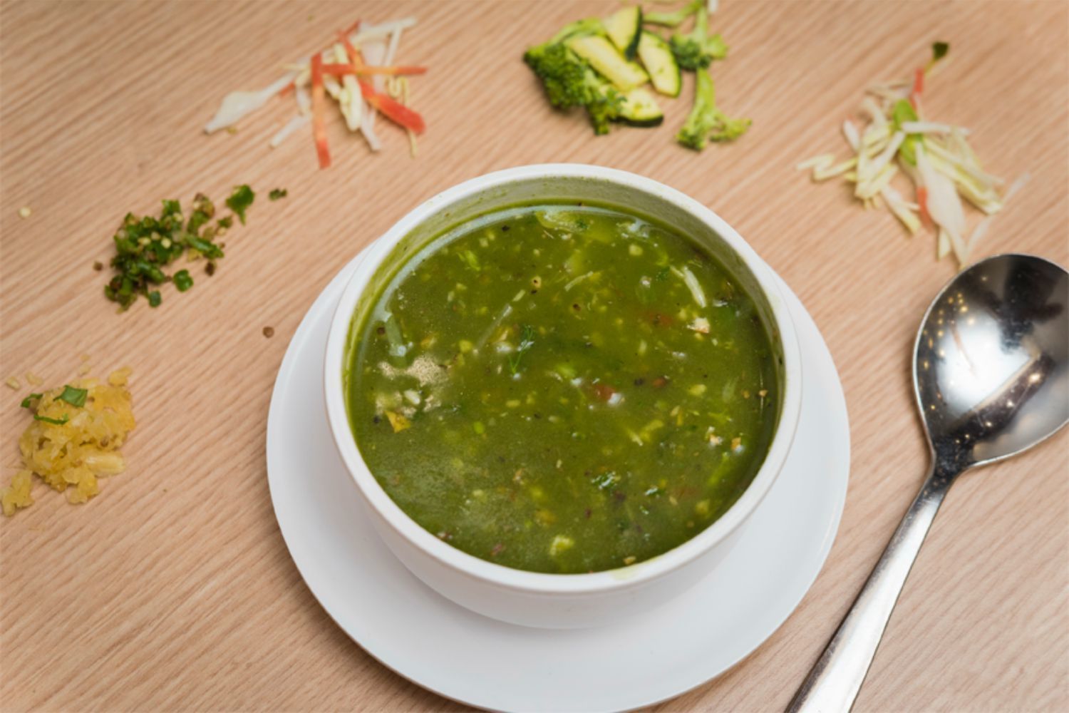 A bowl of soup on a white plate with a spoon beside it, surrounded by vegetable scraps on a wooden table.