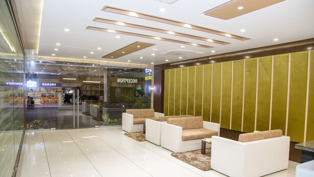 A modern lobby with a reception desk, seating area, and a bakery visible through the glass doors. - Hotel Orange International (Surat)