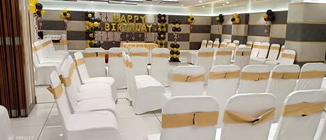 Un salón de banquetes con filas de sillas blancas adornadas con cintas doradas, un escenario con una decoración de globos que dice 'FELIZ CUMPLEAÑOS' y un cartel que dice 'FELIZ CUMPLEAÑOS'.