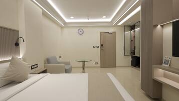 A modern hotel room with a bed, a desk, a chair, a small table, a clock, and a door.