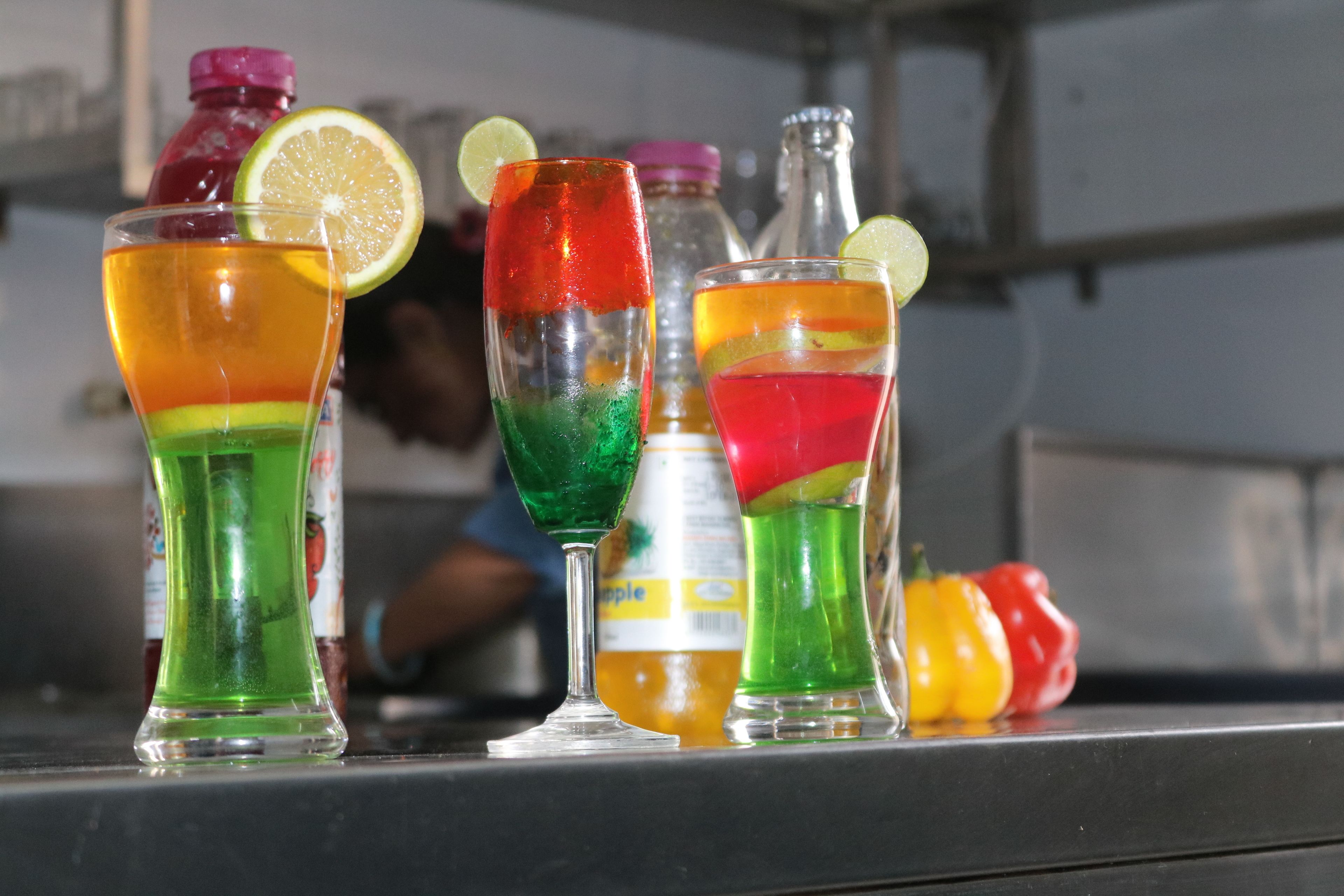A bar area with colorful drinks in glasses, a lime slice garnish, and a bottle of juice in the background.