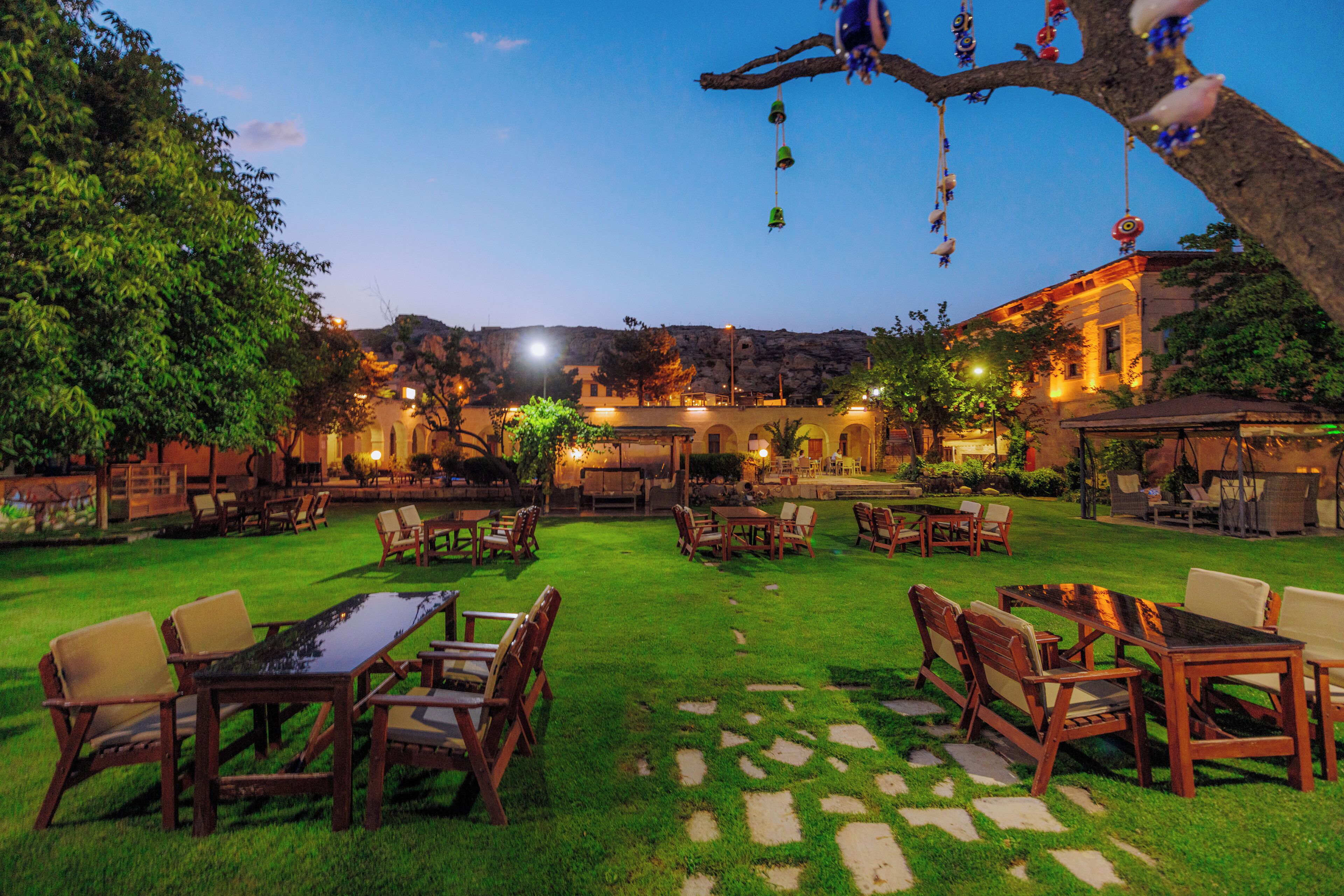 An outdoor patio area with tables and chairs, surrounded by greenery and a building in the background.