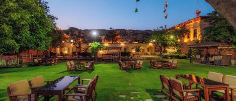 An outdoor patio area with tables and chairs, surrounded by greenery and a building in the background.