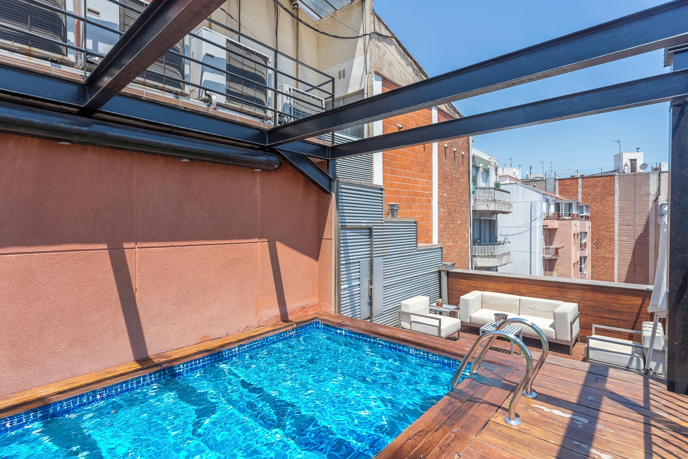 A rooftop pool with a wooden deck, white outdoor furniture, and a view of surrounding buildings.