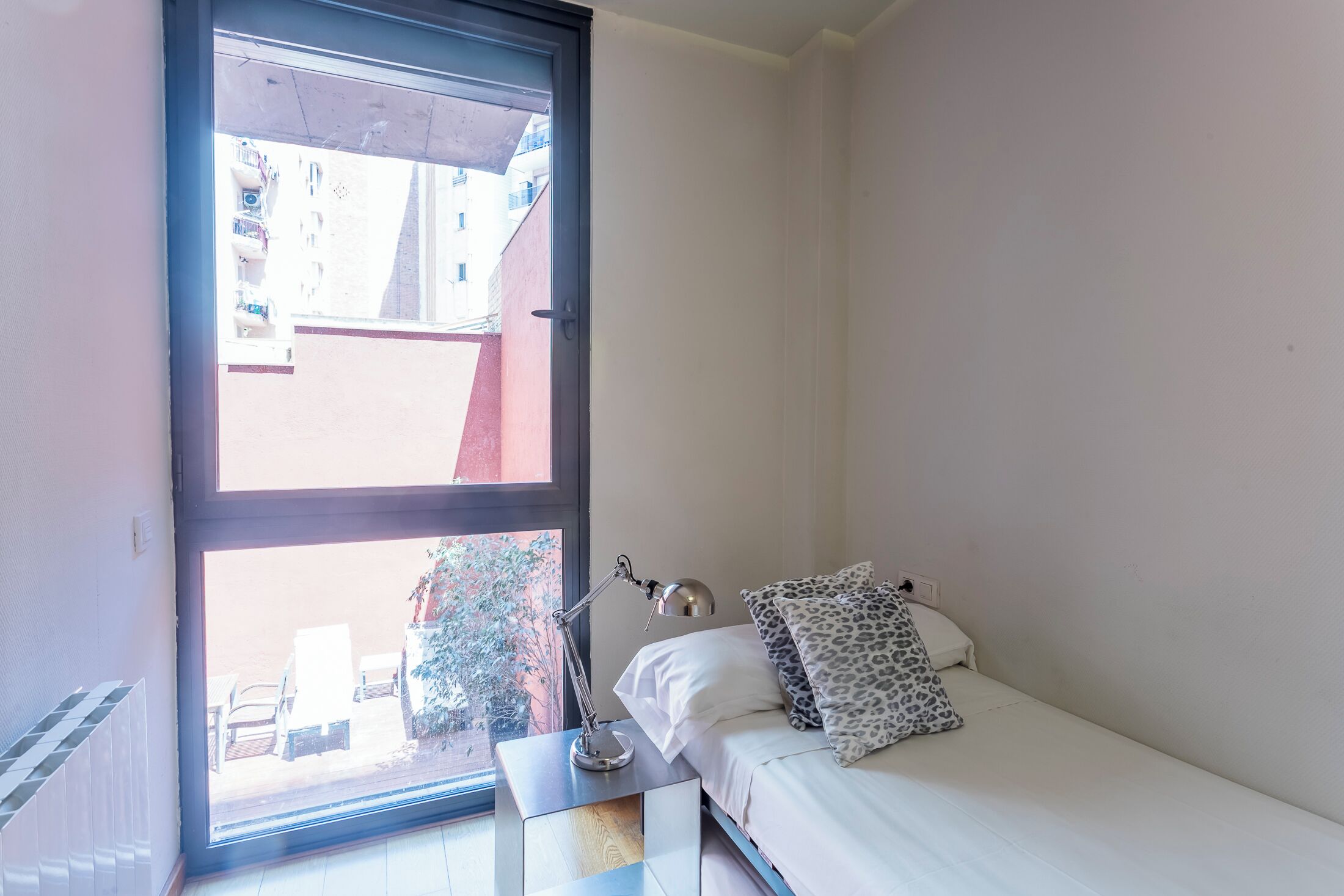 A small, bright room with a bed, a nightstand, a desk, and a window overlooking a balcony.