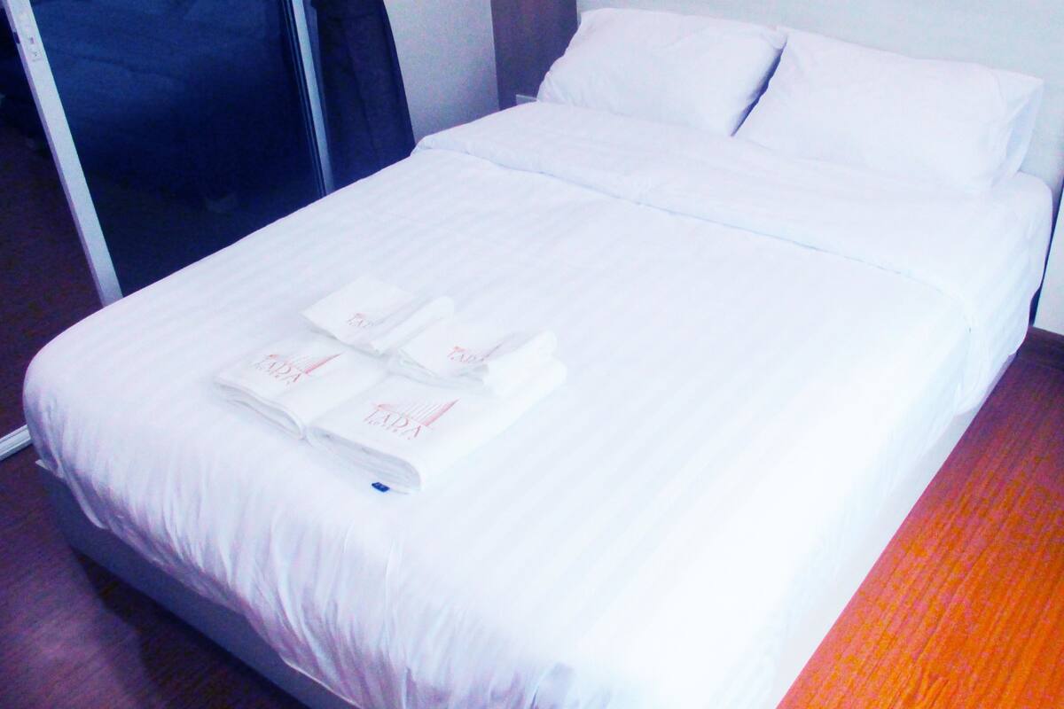 A neatly made bed with white linens and towels, a wooden floor, and a dark-colored piece of furniture to the left.