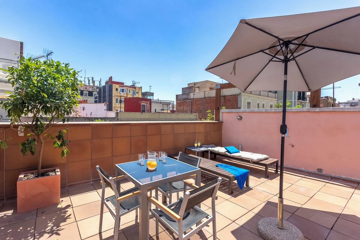 A rooftop terrace with a table, chairs, a large umbrella, and a small tree.
