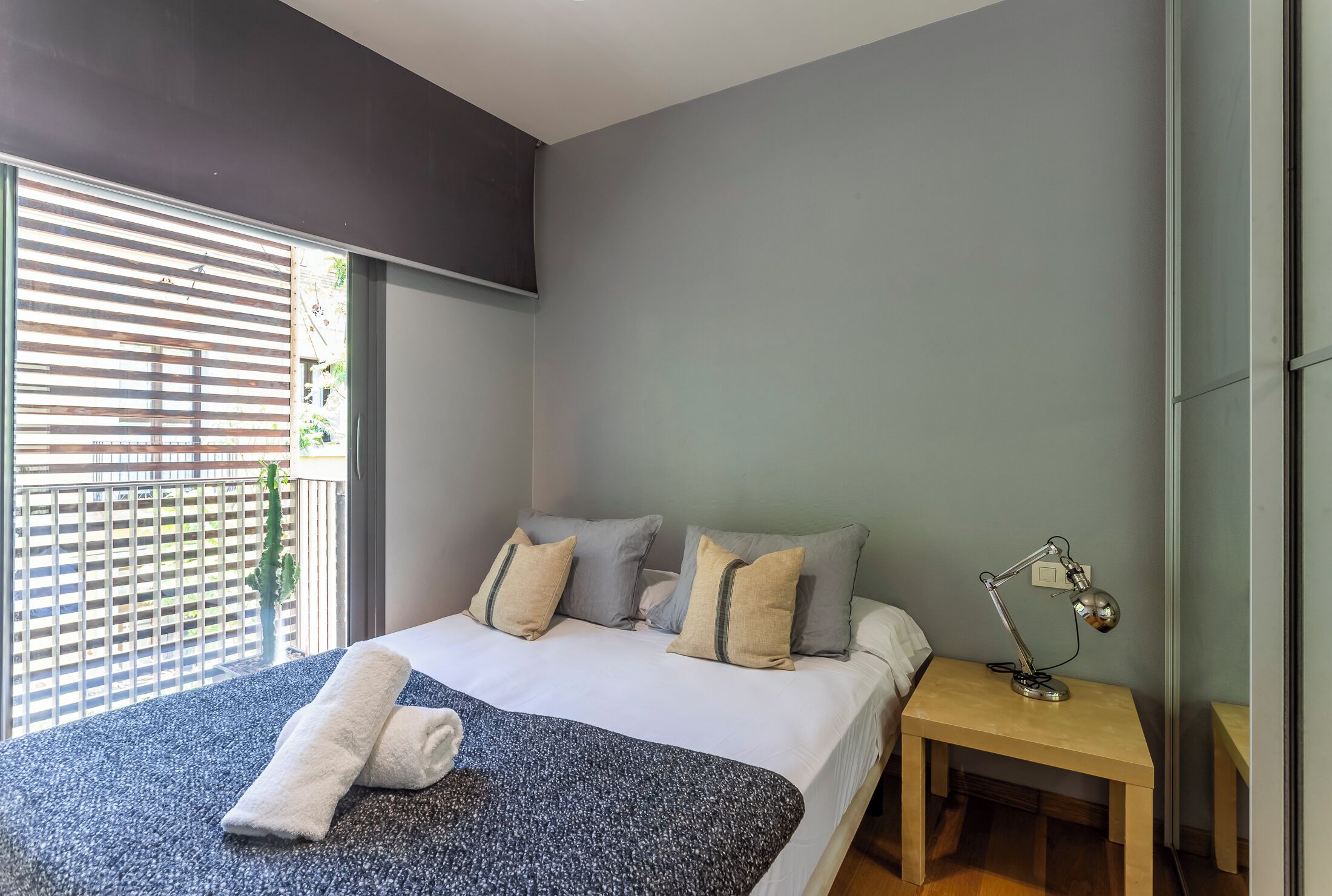 Un dormitorio moderno con una cama grande, una mesita de noche con lámpara y vistas al exterior a través de una ventana con persianas.