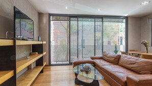 A modern living room with a leather sofa, a glass coffee table, wooden shelves, and a large sliding glass door with blinds.