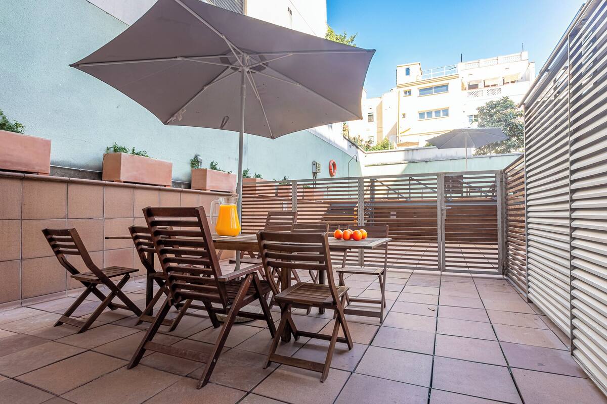 An outdoor patio with wooden chairs and tables, a large umbrella, and a view of buildings in the background.