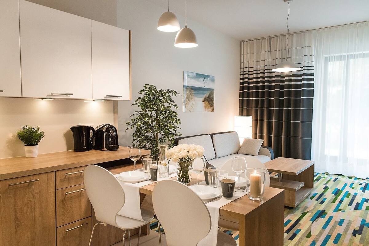 A modern kitchen with white cabinets, a dining area with a wooden table and chairs, a coffee machine, and a potted plant.