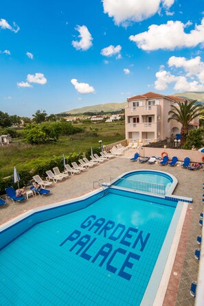 Ein Swimmingpool mit dem Schriftzug „GARDEN PALACE“ auf den Fliesen, umgeben von Liegestühlen und Sonnenschirmen, mit einem Gebäude im Hintergrund.