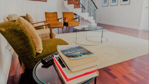 A living room with a green armchair, a stack of books, and a remote control on a glass table. A staircase with a railing is visible in the background. - Comercial Boutique Azores (Ponta Delgada)