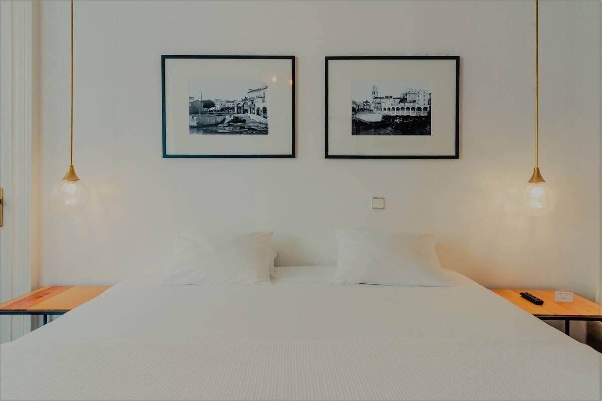 En pænt redt seng med hvide sengetøj, to indrammede sort-hvide fotografier på væggen, og et natbord med en fjernbetjening.