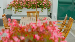 A wooden table with four chairs, a bench, and a planter with pink flowers. - Comercial Boutique Azores (Ponta Delgada)