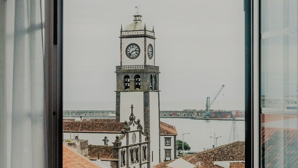 A view from a window overlooking a historic building with a clock tower, a waterfront, and a crane in the distance. - Comercial Boutique Azores (Ponta Delgada)