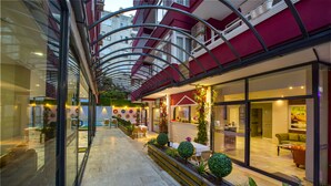 A modern hotel corridor with glass walls, a covered outdoor seating area, and a visible restaurant. - Twin Apart Hotel (Alanya)