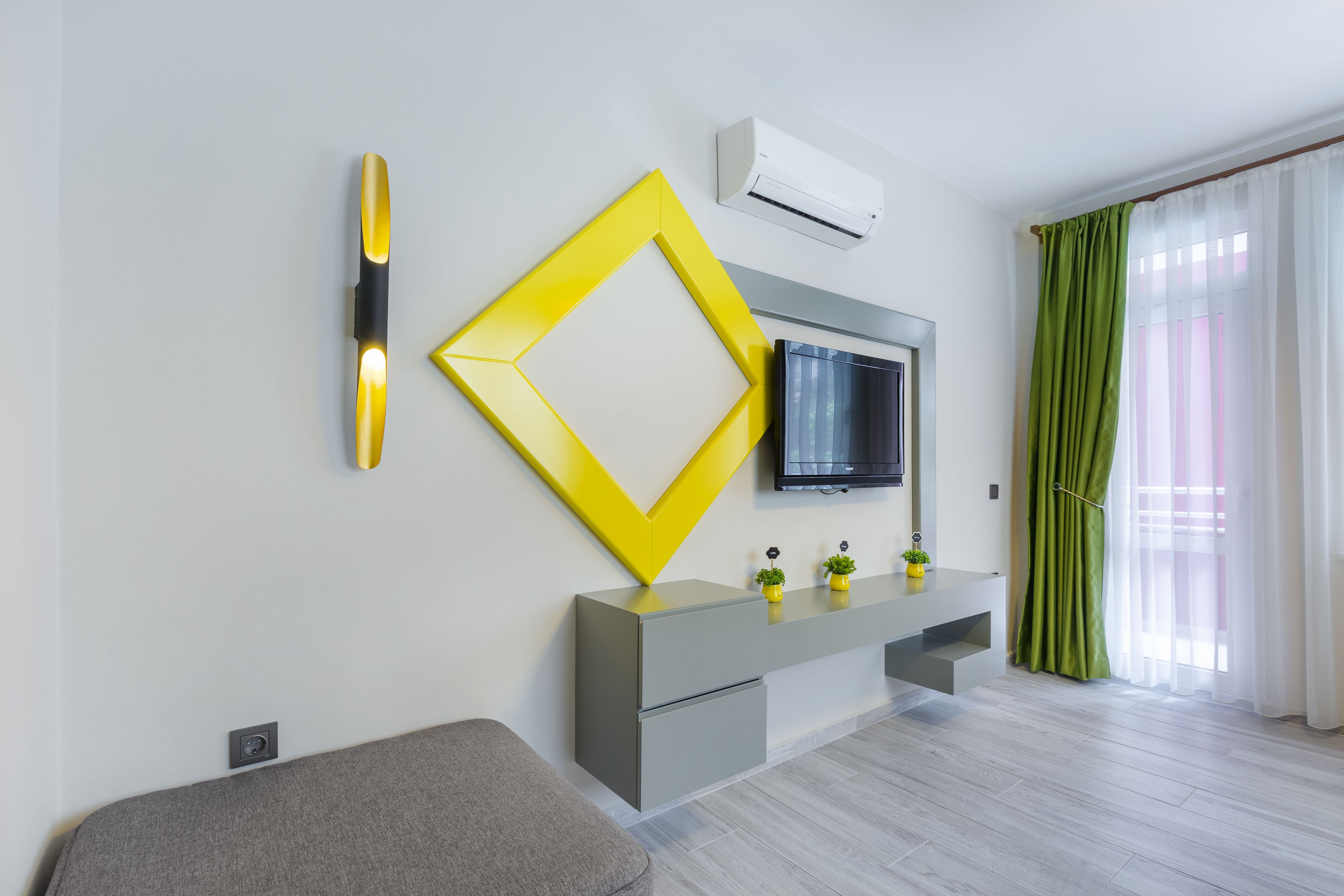 Una sala moderna con un sofá gris, un adorno amarillo, una mesa consola gris, un televisor de pantalla plana y una cortina verde.