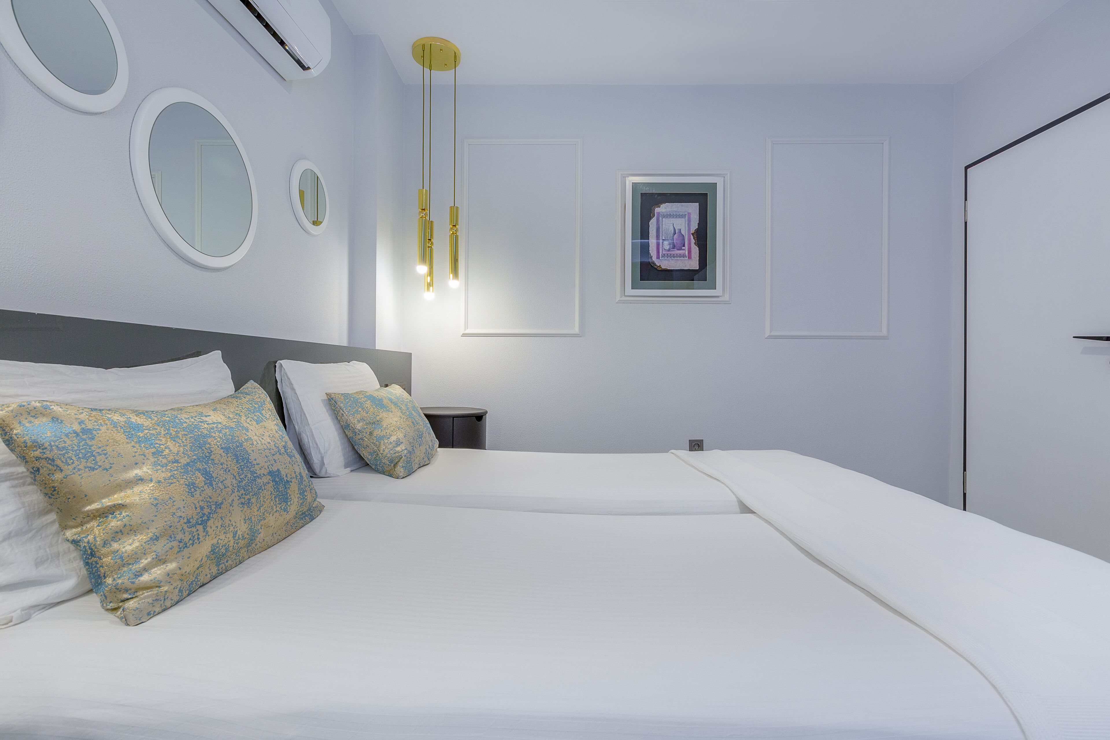 A modern hotel room with a large bed, two bedside tables, a wall-mounted air conditioner, and a ceiling light fixture.