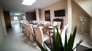Un comedor con una mesa larga dispuesta para seis personas, rodeada por seis sillas. Hay un televisor montado en la pared y un ventilador de pie cerca de la mesa.