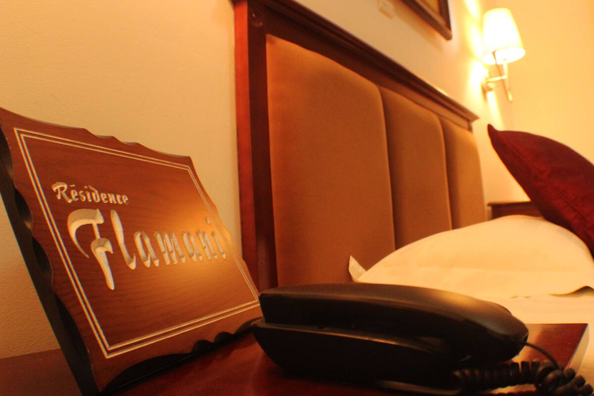 A hotel room with a wooden headboard, a bedside table with a telephone, a pillow, and a wall-mounted lamp.