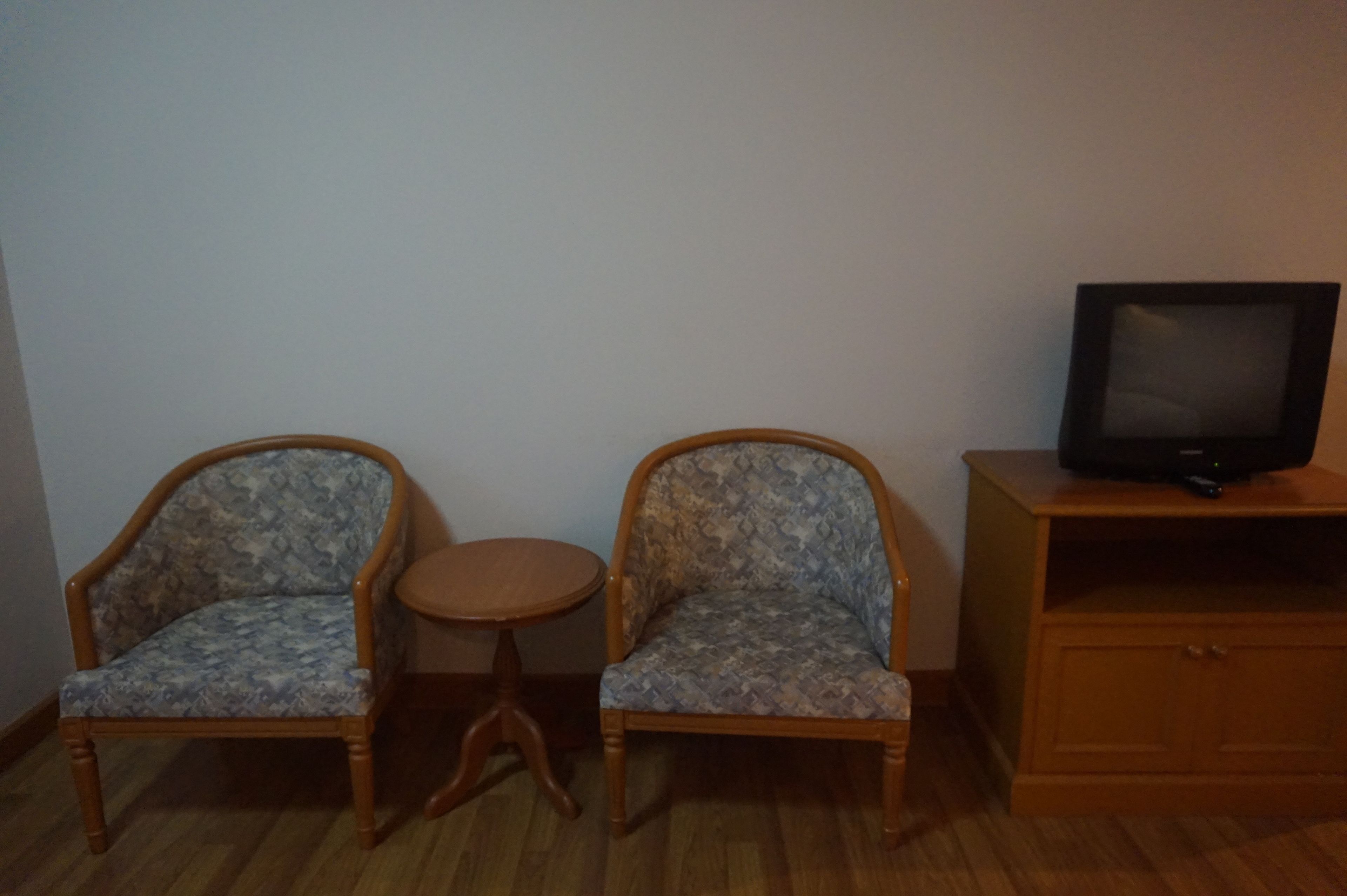 Ein Zimmer mit zwei gemusterten Sesseln, einem kleinen runden Tisch, einem Fernseher auf einem Holzgestell und einem Schrank.