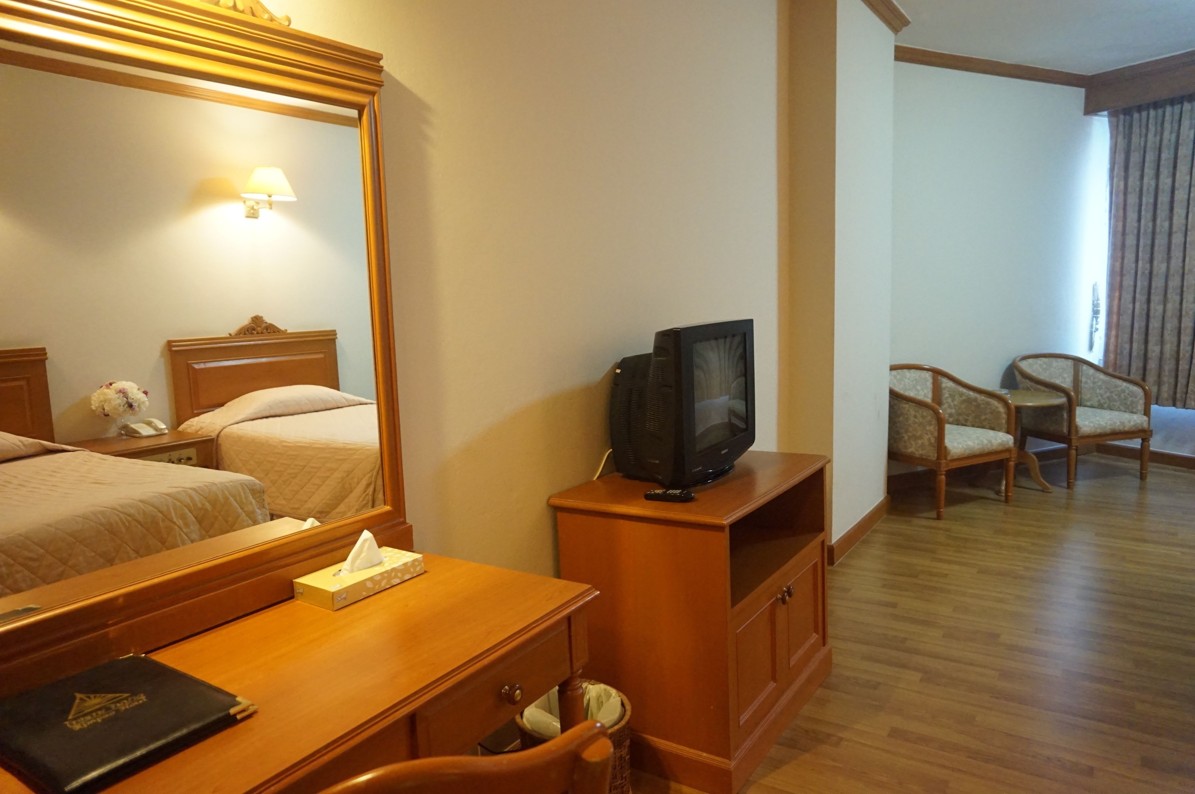 A hotel room with a wooden desk, a television, a mirror, two beds, a chair, and a window with curtains.