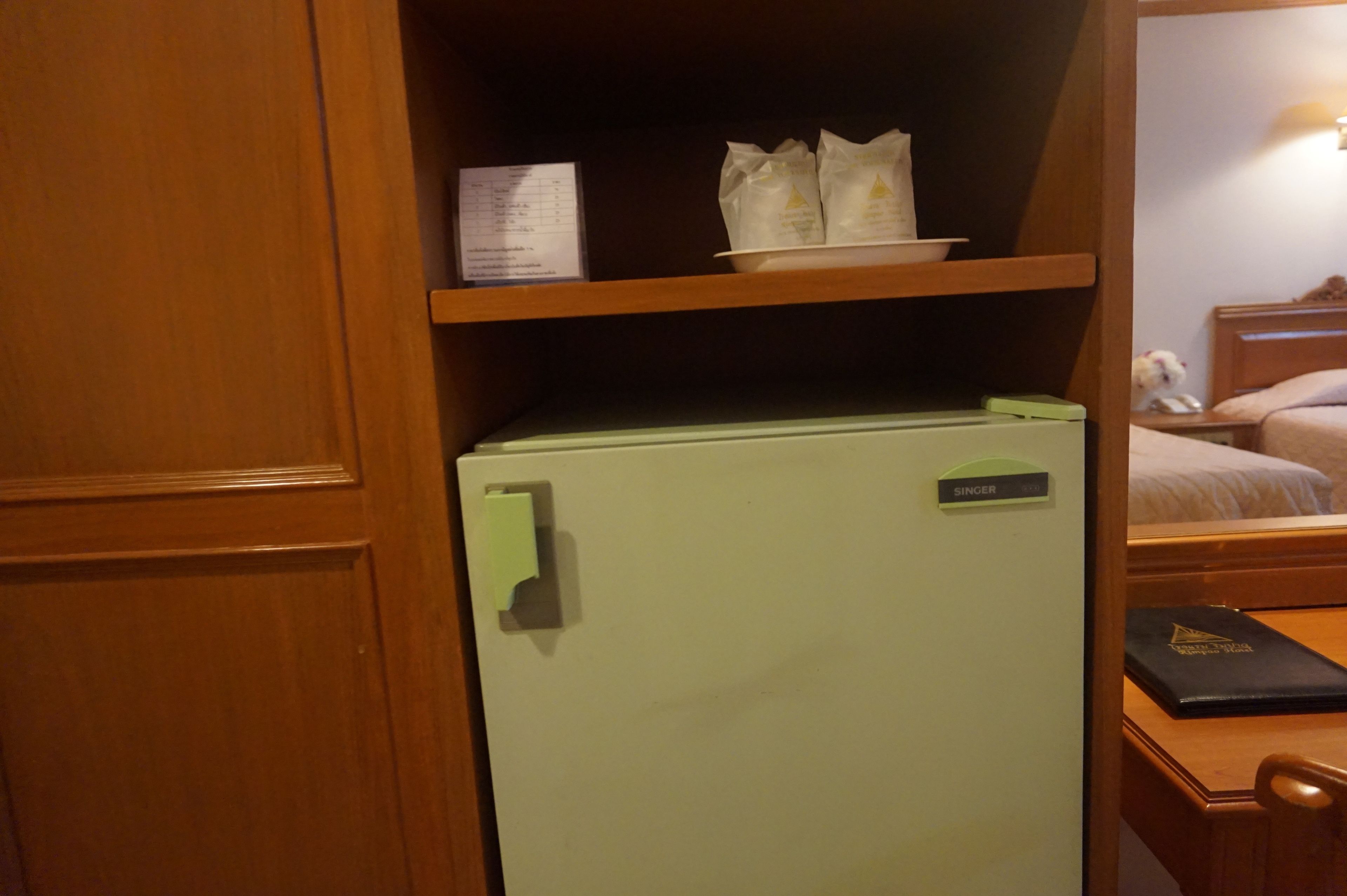 A hotel room with a green minibar, a nameplate reading "SINGER", a small shelf with a menu, and a bed with a nightstand.