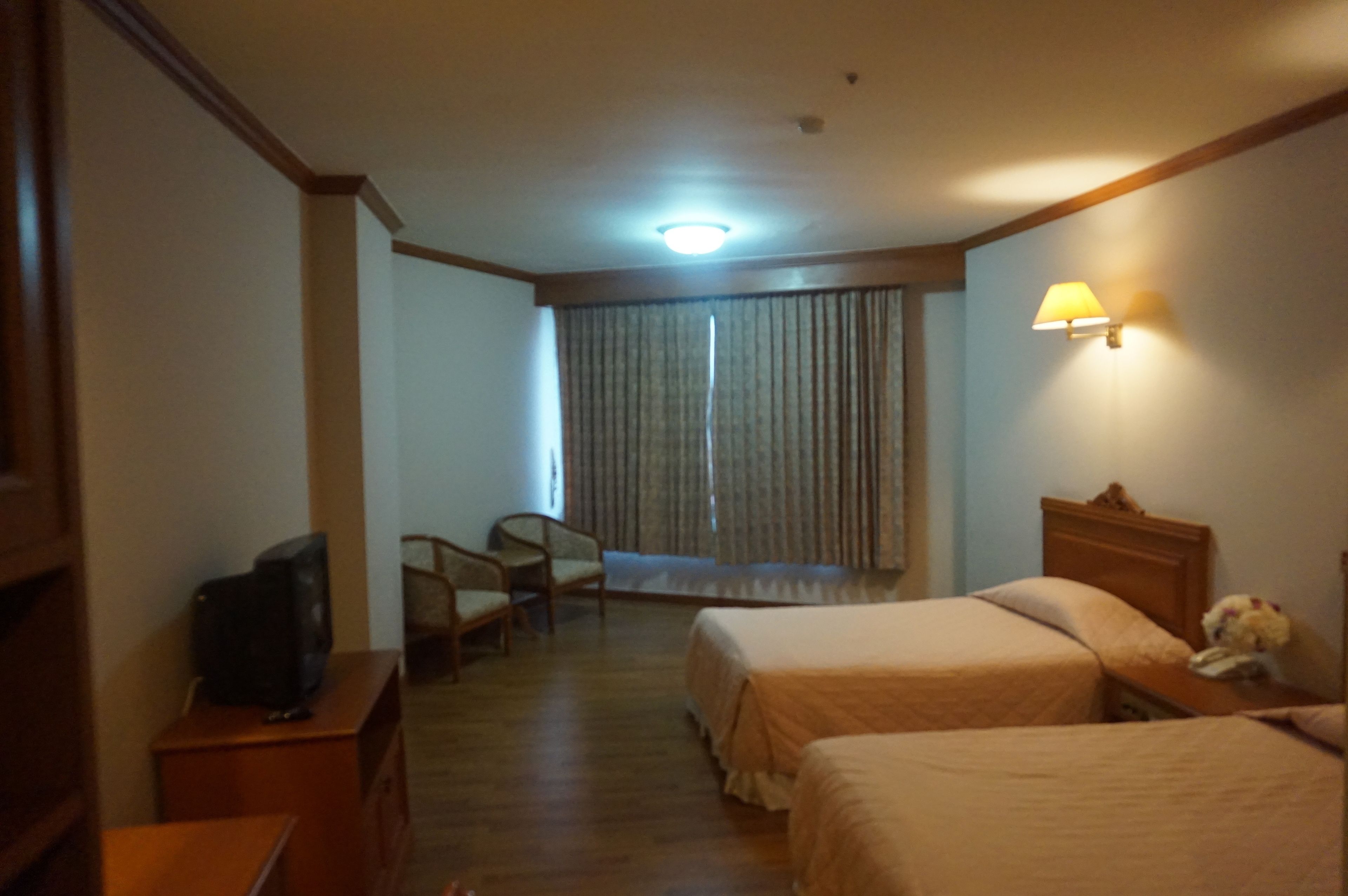 A hotel room with two beds, a television on a side table, a lamp on the wall, and a window with curtains.