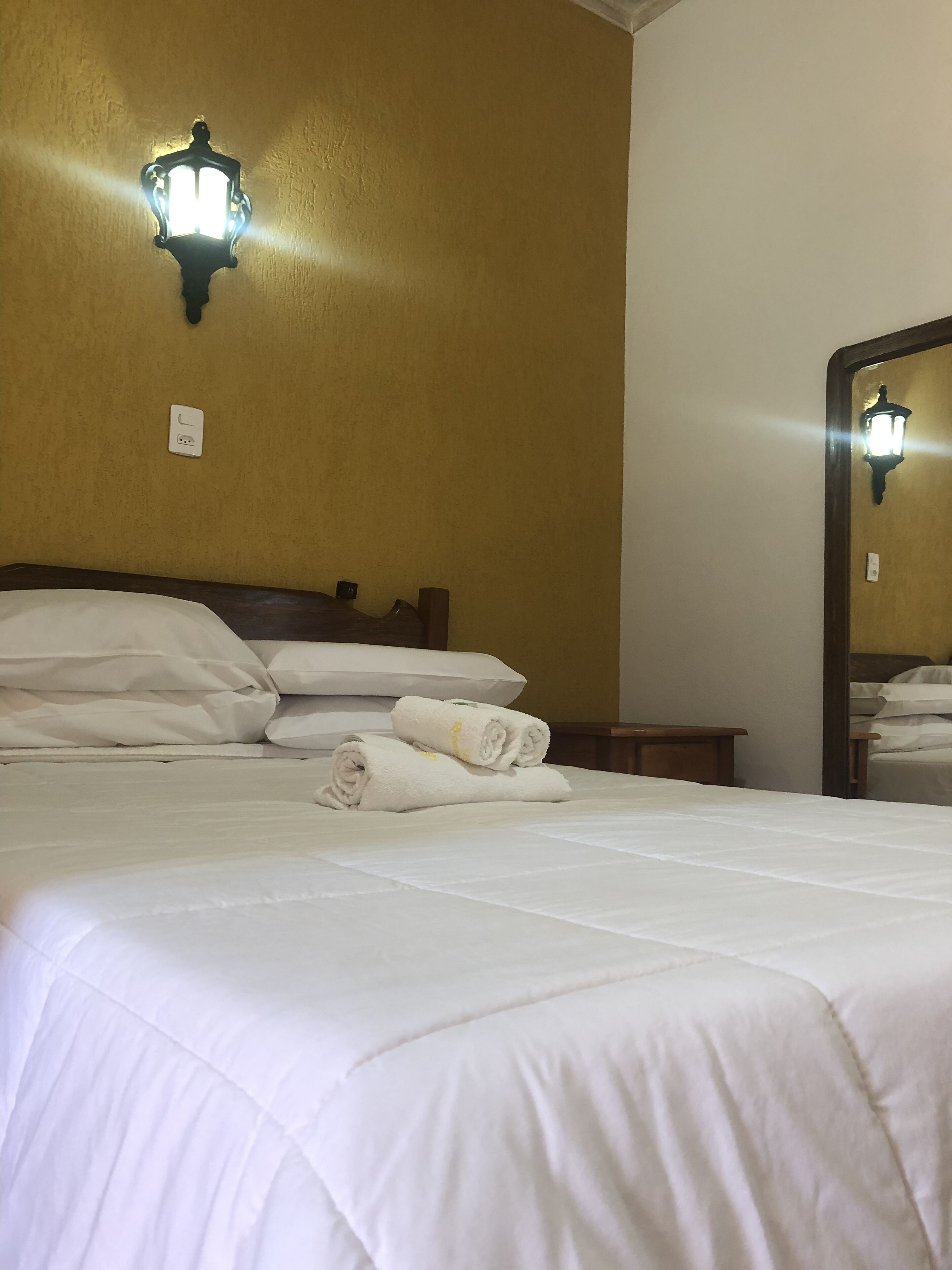 A neatly made bed with white linens, a wall-mounted light fixture, and a mirror reflecting part of the room.