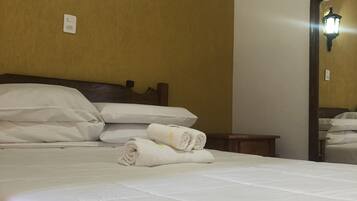 A neatly made bed with white linens, a wall-mounted light fixture, and a mirror reflecting part of the room.