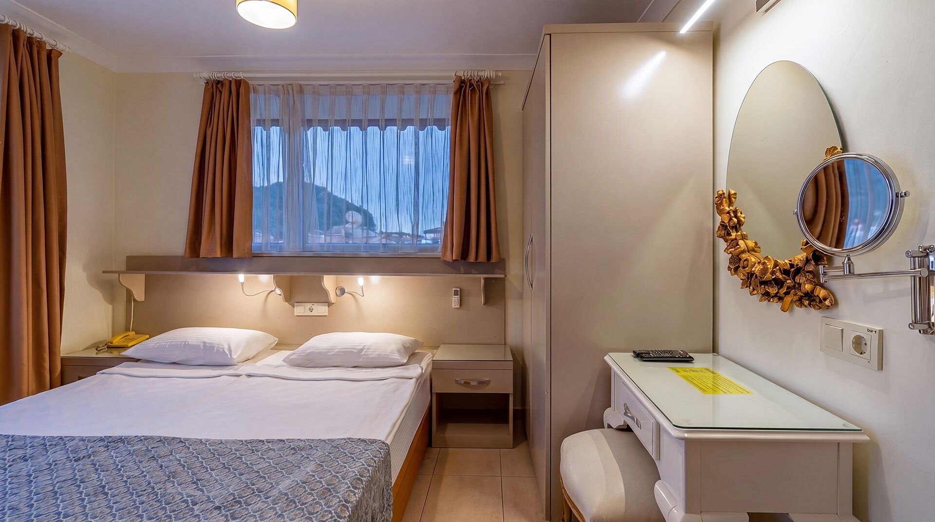 A hotel room with a bed, bedside table, lamp, mirror, and a desk with a chair.